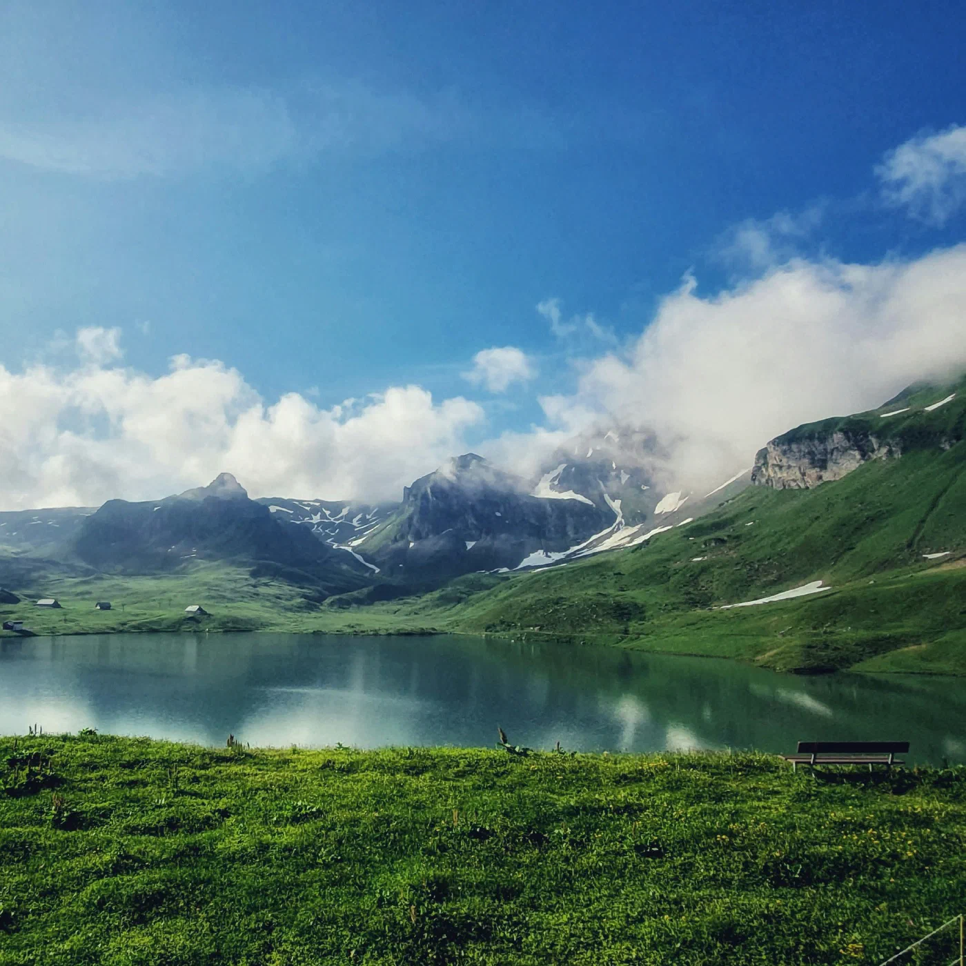 A calm alpine lake reflecting green hillsides and cloud-wrapped peaks